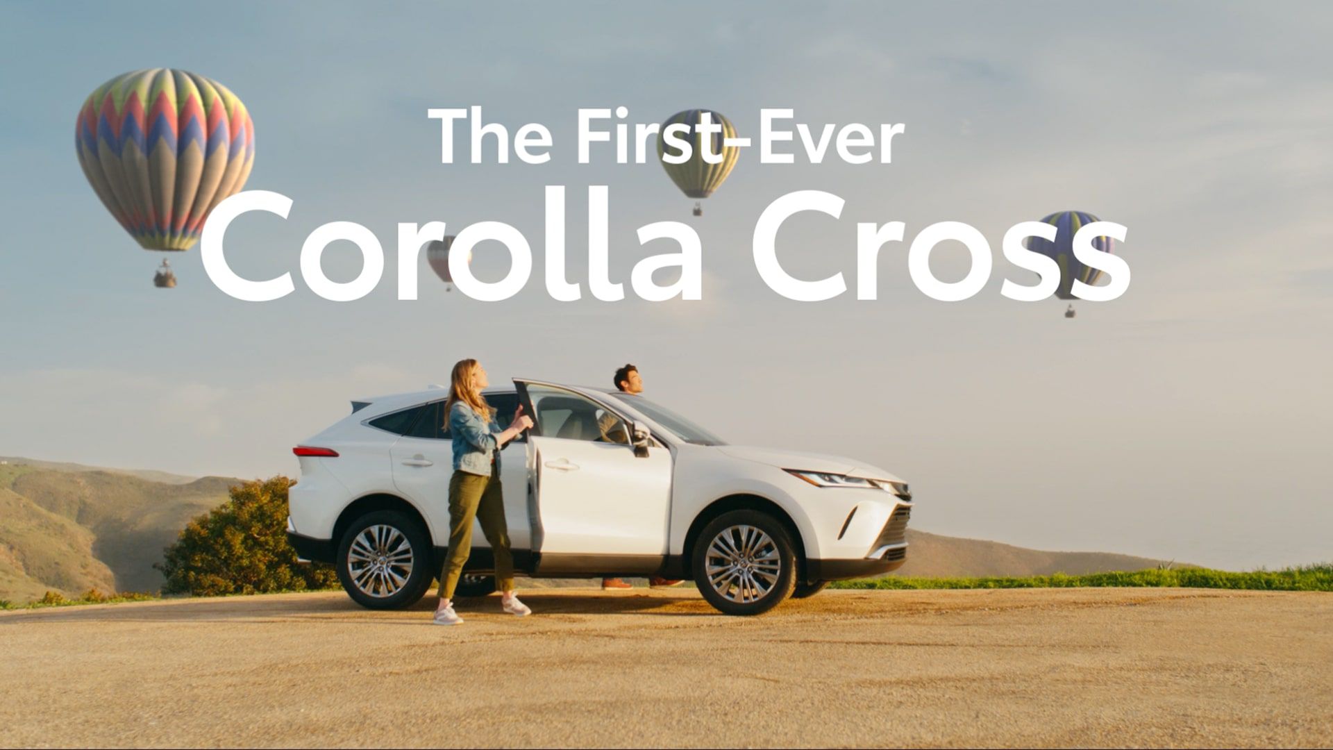 A couple gets out of a vehicle to look at hot-air balloons in the sky. Text across the image reads “The First-Ever Corolla Cross.”