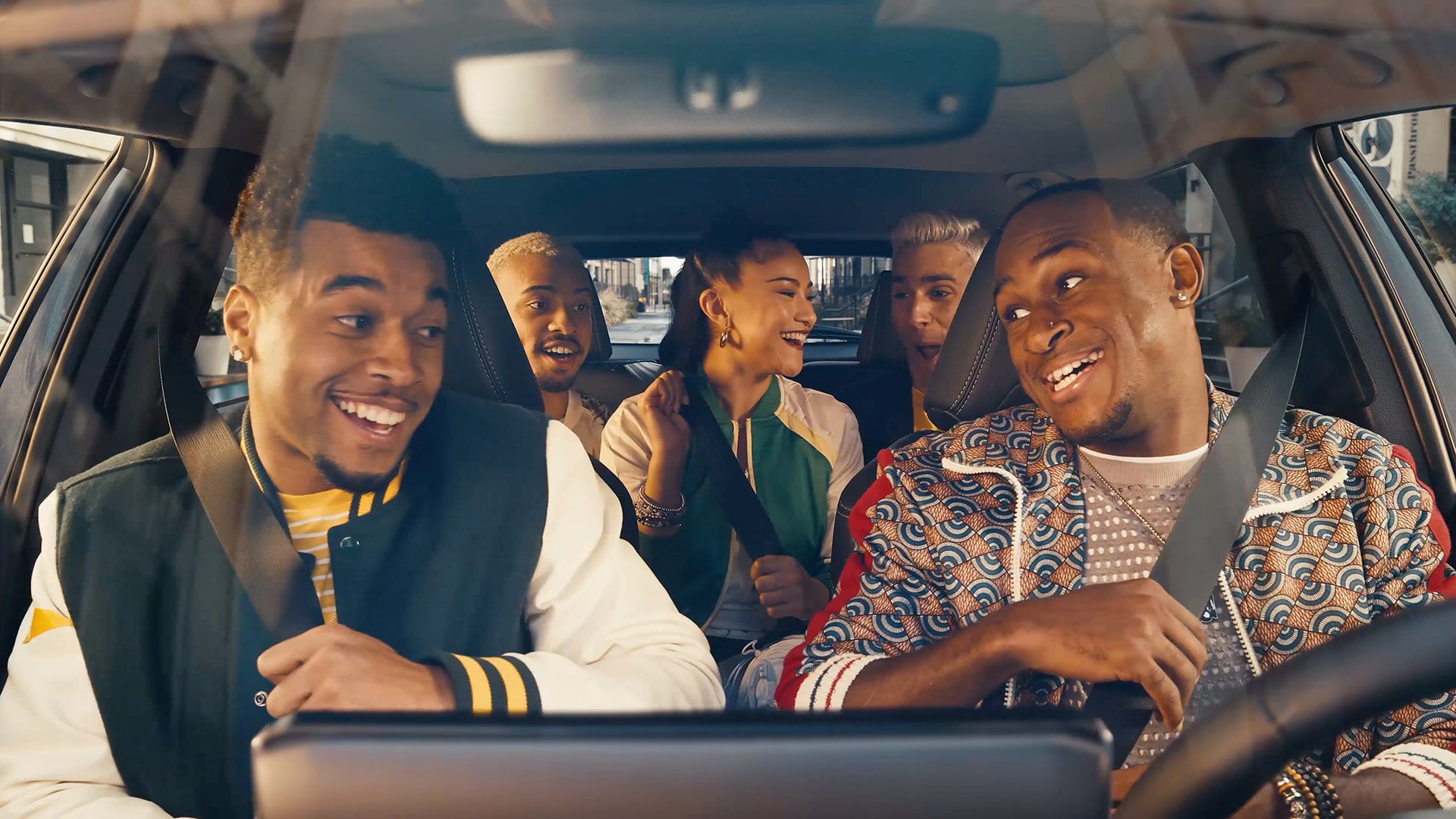 Two friends in front and three friends in the back seats are all smiling at one another as they put on their seatbelts.