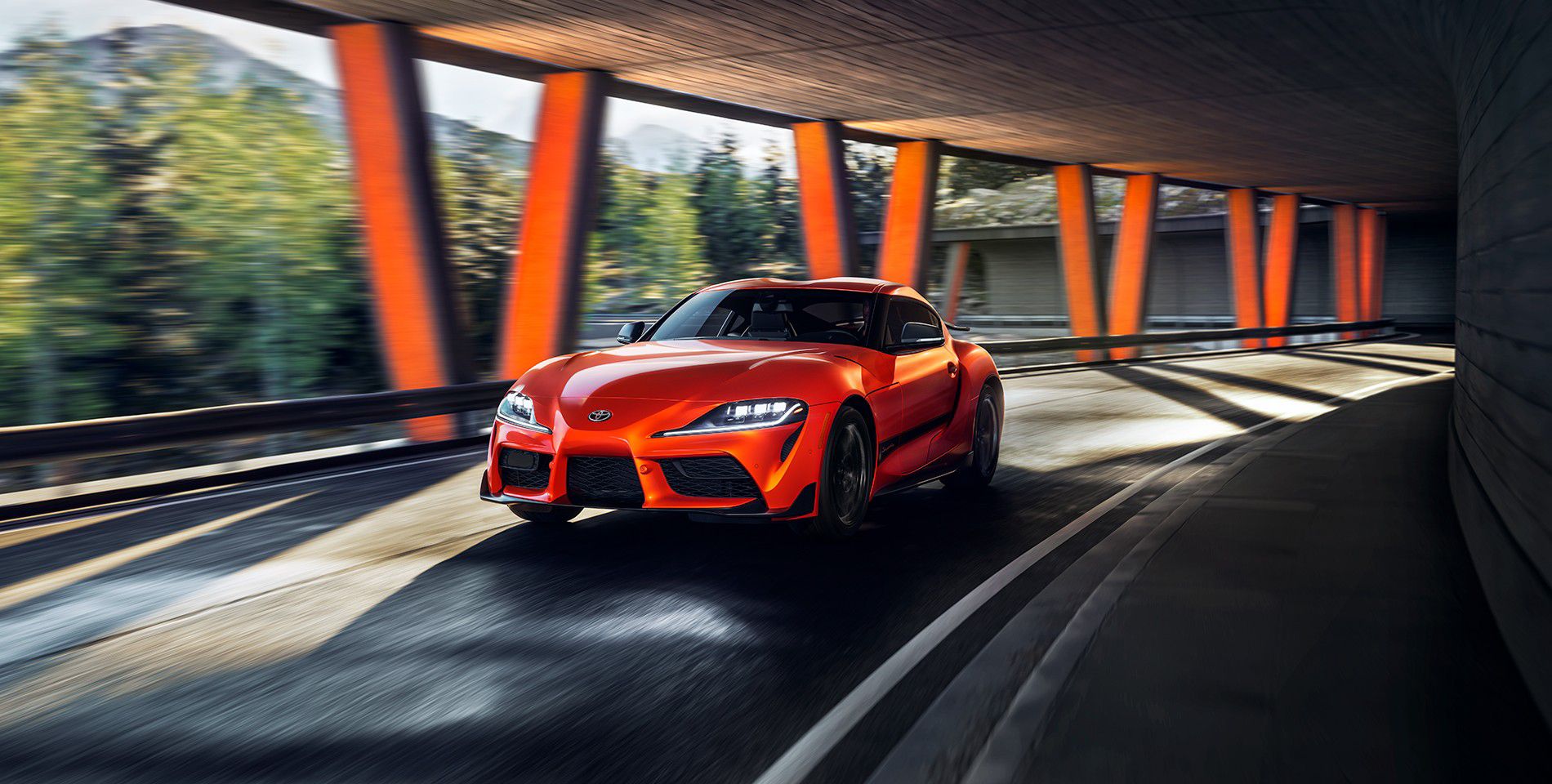 A red Toyota vehicle turns along a curving road underneath a covered bridge.