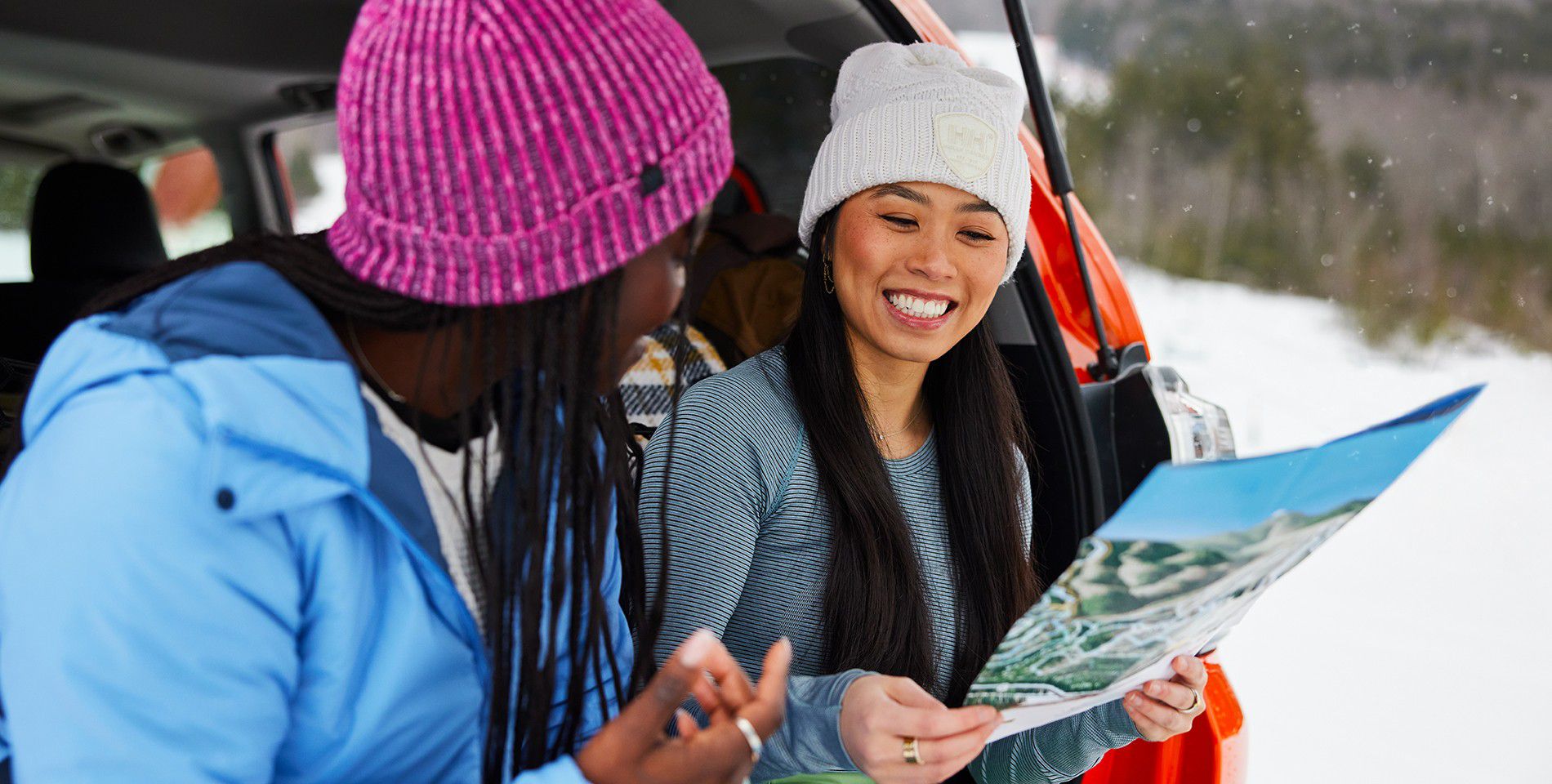 Two girls wearing winter beanies in a snowy landscapre lean against the back of a Toyota vehicle looking at a trail map and smiling.