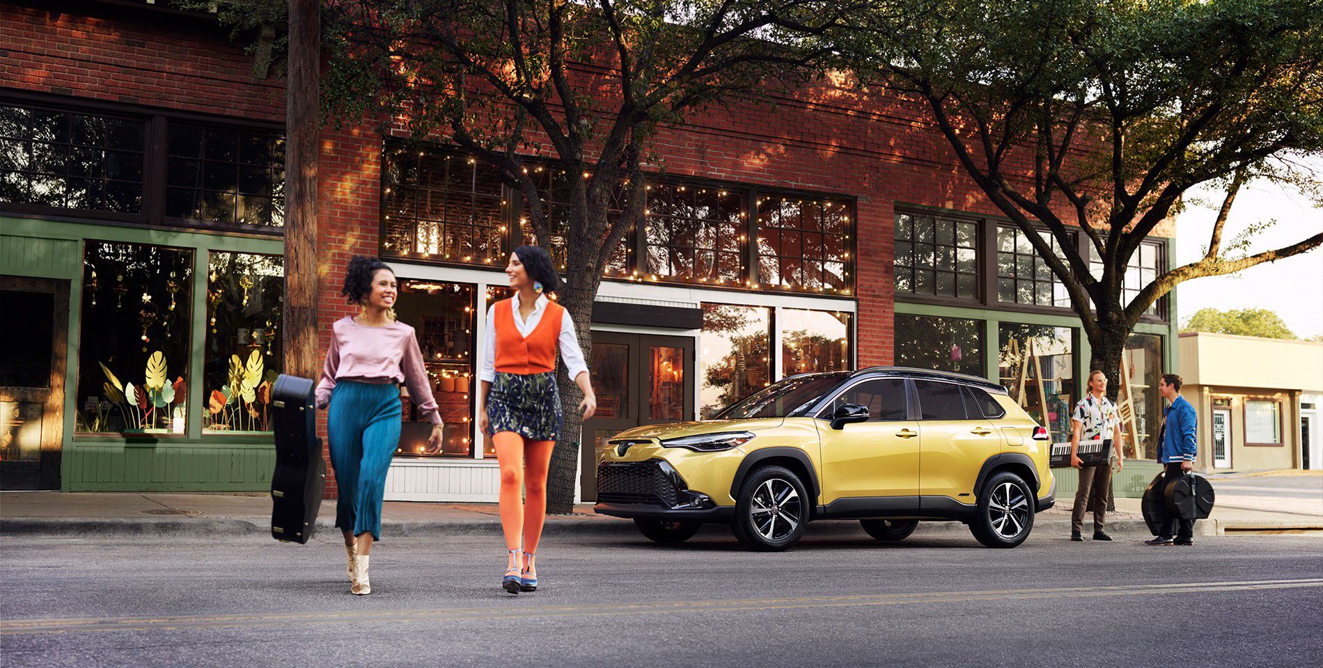 Two girls wearing bright clothing cross a shady street with stores and a bright yellow Toyota vehicle in the background.