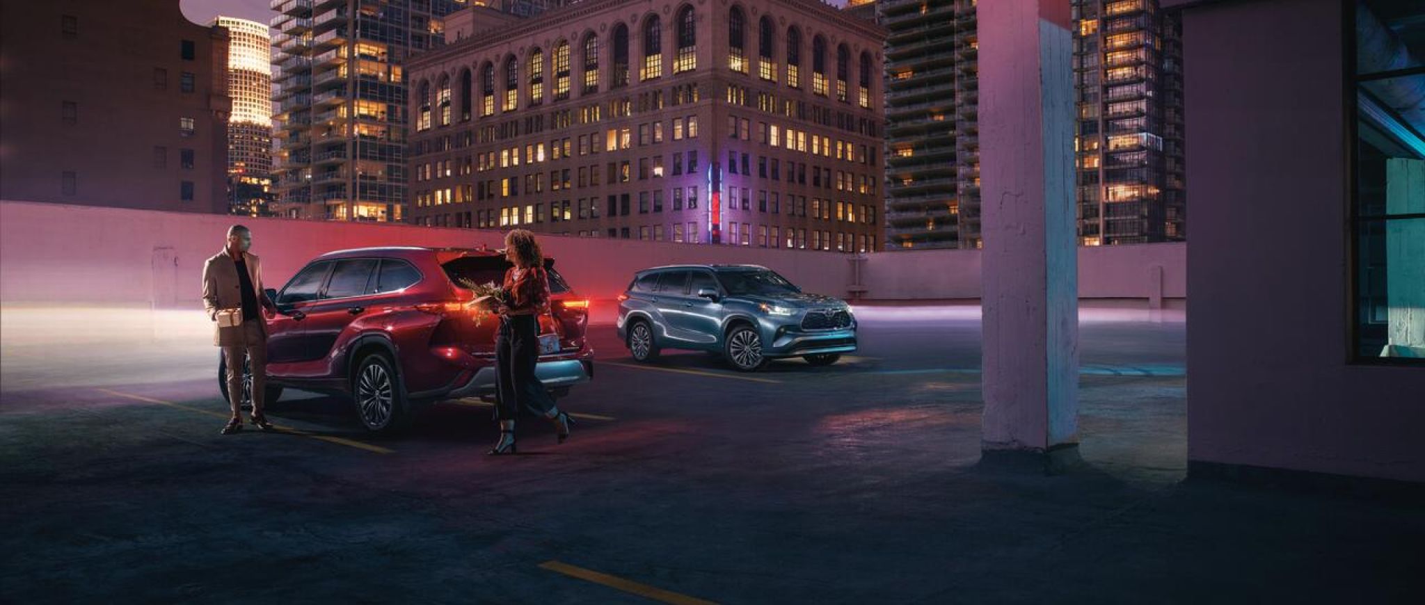 A night-time view of the sleek body lines of a Ruby Flare Pearl colored and a Stone Blue colored Toyota Highlander SUV. 
