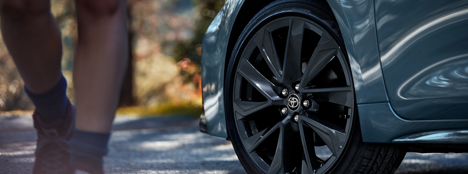 La rueda delantera de un Toyota Camry muestra sus llantas de aleación y el perfil delantero derecho sobre un fondo borroso de bosque; por el lado pasa caminando una persona en pantalones cortos.