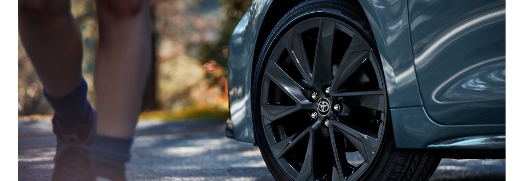 La rueda delantera de un Toyota Camry muestra sus llantas de aleación y el perfil delantero derecho sobre un fondo borroso de bosque; por el lado pasa caminando una persona en pantalones cortos.