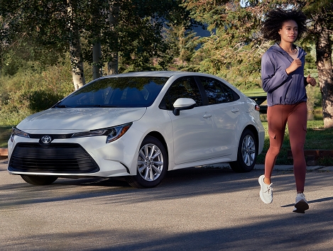 Un Toyota Corolla Hybrid en color blanco estacionado detrás de  arbustos verdes. Por su lado pasa corriendo una joven con zapatillas deportivas blancas, pantalones de yoga color granate y una camisa de poliéster.