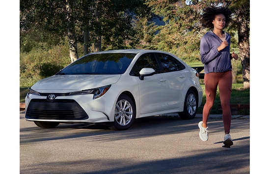 A Toyota Corolla Hybrid in white is parked behind verdant green bushes. A young lady in white sneakers, maroon yoga pants, and polyester shirt jogs by.