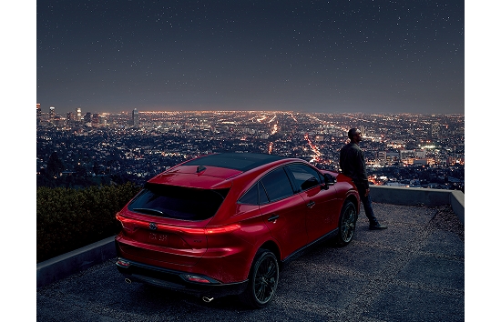 A 2023 Toyota Venza in the color Barcelona Red Metallic sits atop a promontory with unhindered view of a city skyline and lights; a man leans against the front of the vehicle.