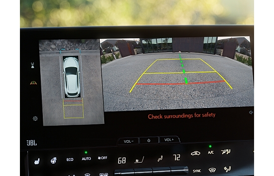 Un Toyota blanco usando la asistencia para estacionar inteligente con líneas de guía a color para ayudar al conductor a estacionar el vehículo en un estacionamiento urbano pavimentado con casas enfrente.