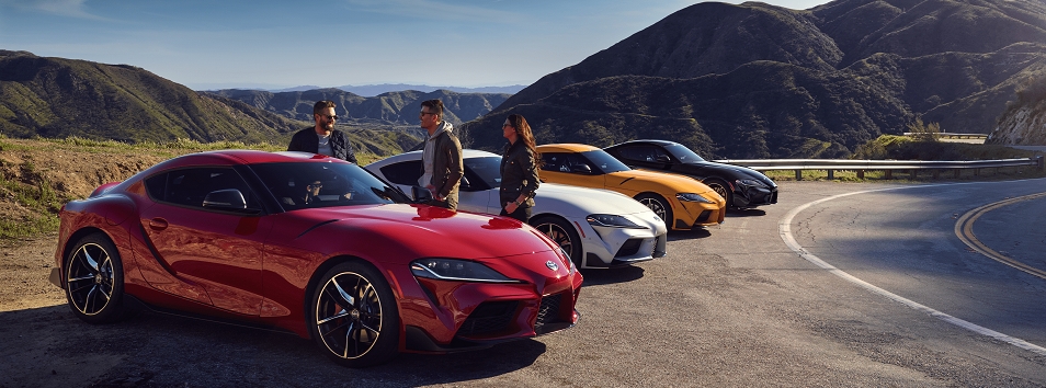 A row of four Toyota Supras in Renaissance Red 2.0, Nitro Yellow, Tungsten, and Nocturnal are parked on the unpaved side of a mountain road. A group of three people gathered around the first car