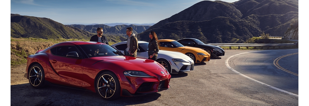Una fila de cuatro Toyota Supra en Renaissance Red 2.0, Nitro Yellow, Tungsten y Nocturnal estacionados en el lado sin pavimentar de una carretera montañosa. Un grupo de tres personas reunidas alrededor del primer auto.