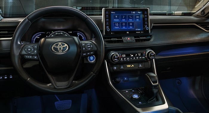 The interior of a 2023 Toyota RAV4 Hybrid in black displaying its steering wheel, instrument cluster, knobs, controls, multimedia and infotainment screen.