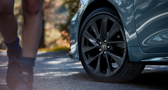The front wheel of a Toyota Camry shows its alloy rims and front right profile against a blurred-out wooded background; a person in shorts walks by.