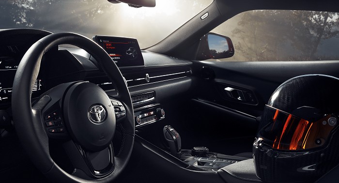 The interior of a 2023 Toyota Supra in black. Pictured are the steering wheel, instrument cluster, shifter, knobs, and controls; buttons with red LED accent lighting