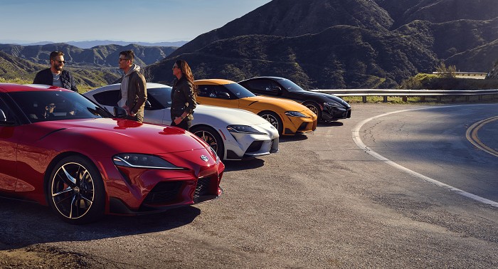 Una fila de cuatro Toyota Supra en Renaissance Red 2.0, Nitro Yellow, Tungsten y Nocturnal estacionados en el lado sin pavimentar de una carretera montañosa. Un grupo de tres personas reunidas alrededor del primer auto.