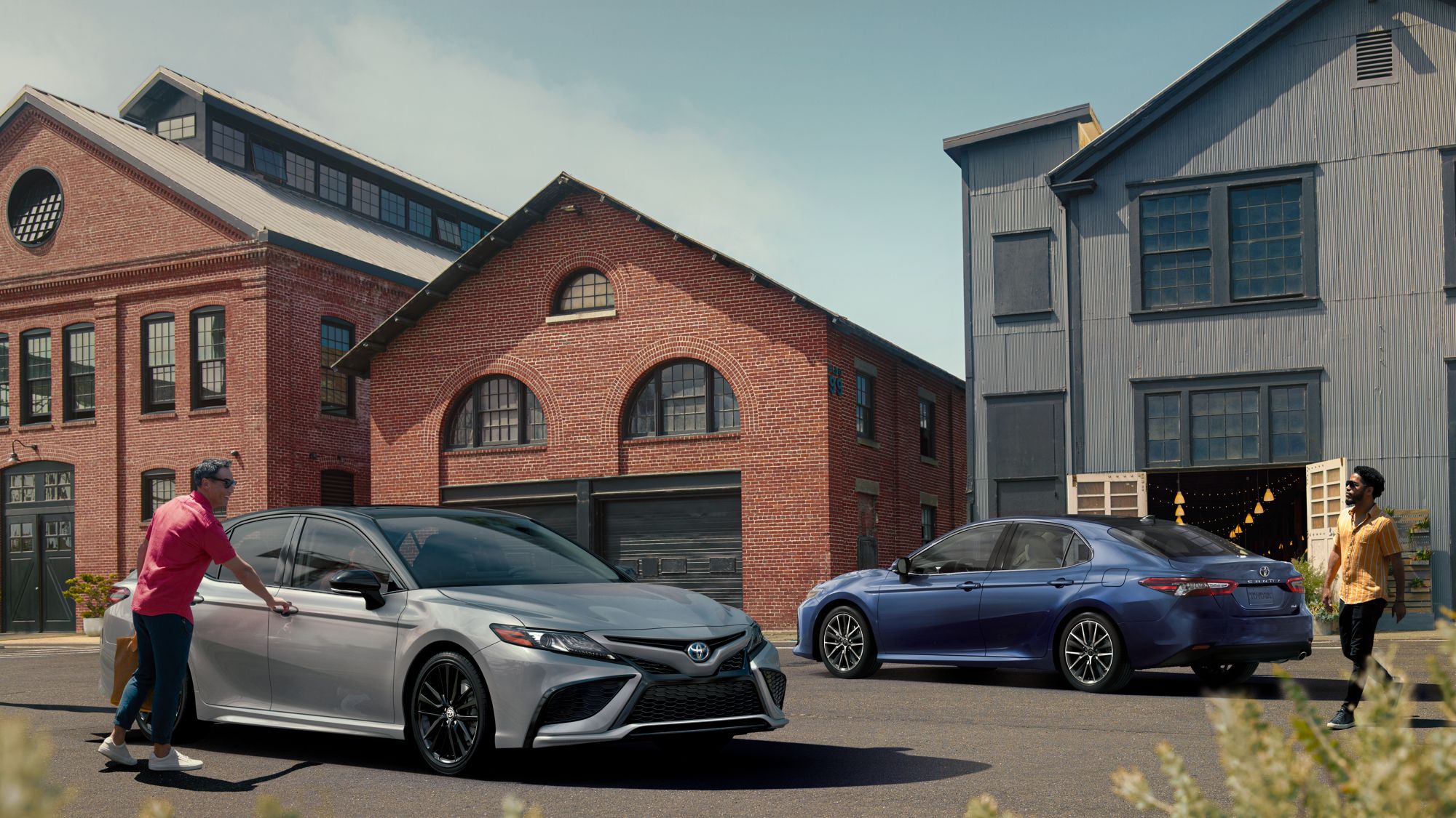 a silver car with a man getting into the from passenger and a blue car parked next to brick buildings. 