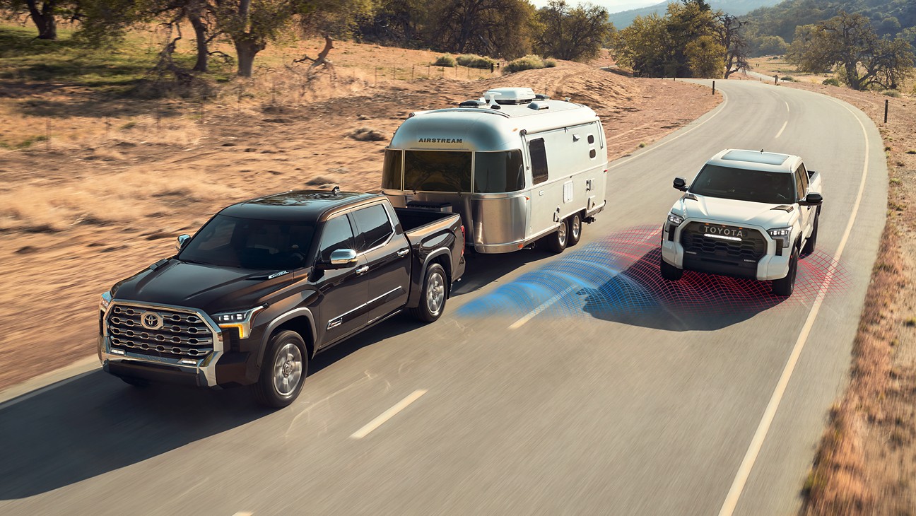 a brown truck hauling a silver camper with a white truck driving next to it. 