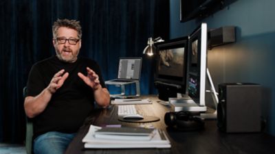 A video editor sits at his work desk. He is talking to the camera as he gestures with his hands.